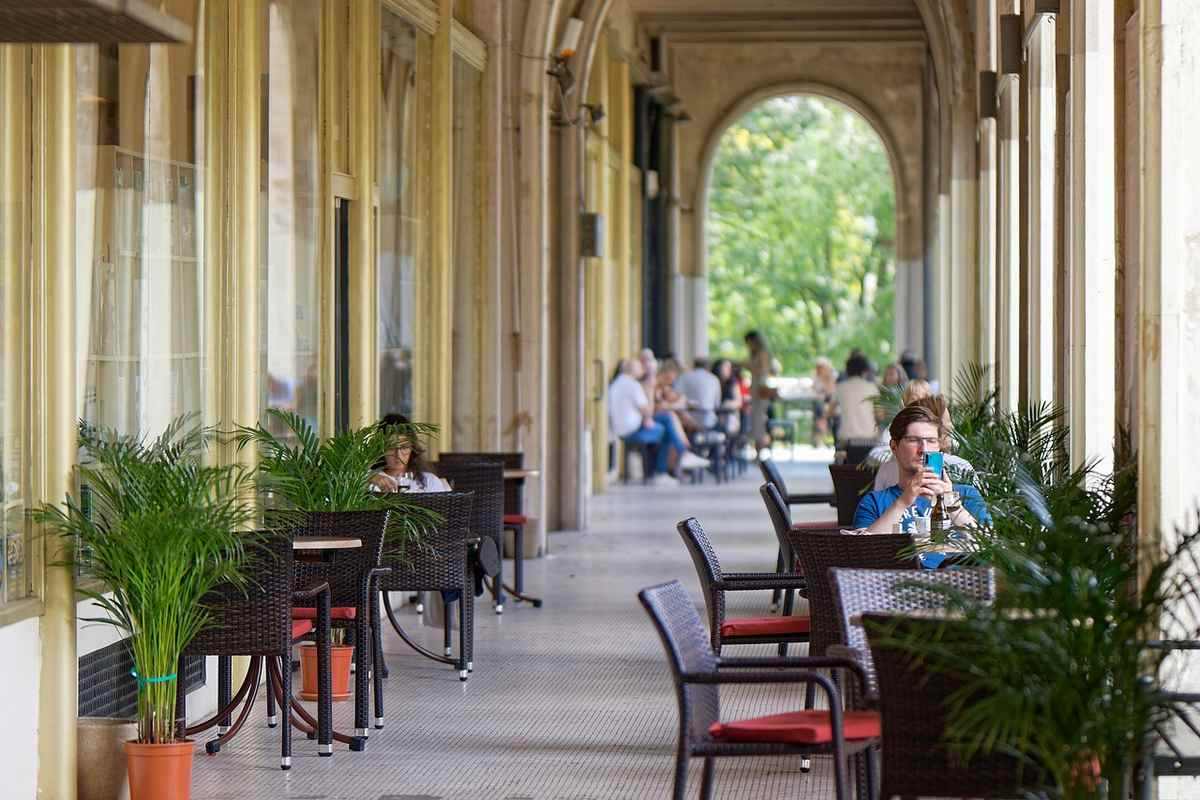 Corridor couvert avec arches, tables et chaises, personnes assises, plantes vertes Corridor couvert avec arches, tables et chaises, personnes assises, plantes vertes