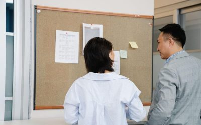Deux personnes regardent un panneau d'affichage en liège avec documents épinglés sur un mur blanc de bureau