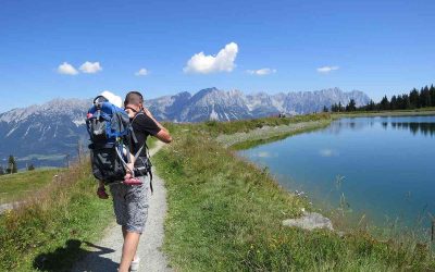 Un randonneur porte son enfant sur le dos, marche près d'un lac de montagne