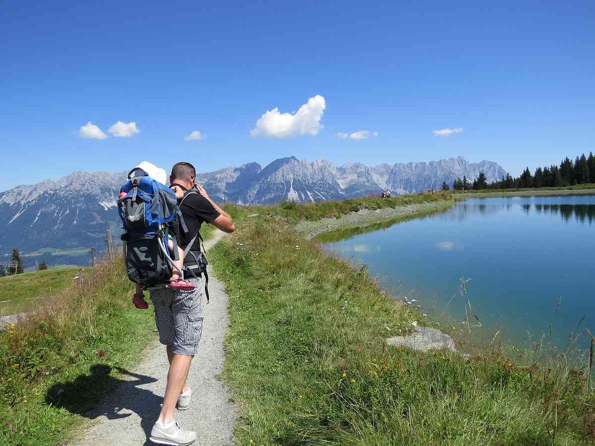 Un randonneur porte son enfant sur le dos, marche près d'un lac de montagne
